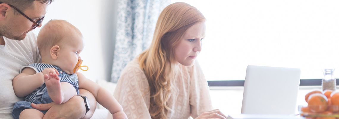 Dad holds their daughter at the kitchen table while Mom checks her email, minus baby, on her laptop next to him.