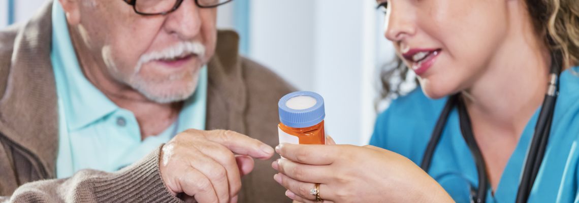 A home caregiver helping a senior man take his prescription medicine. She is holding the pill bottle she is giving him instructions, helping him read the label. The nurse is an Hispanic woman in her 30s and the patient, also Hispanic, is in his 80s. The focus is on their hands and the bottle.