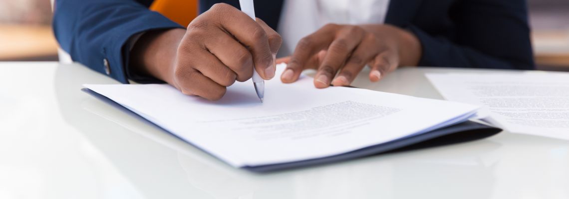 Businesswoman signing contract. African American business woman sitting at table in office, holding pen and writing in document. Legal expertise concept