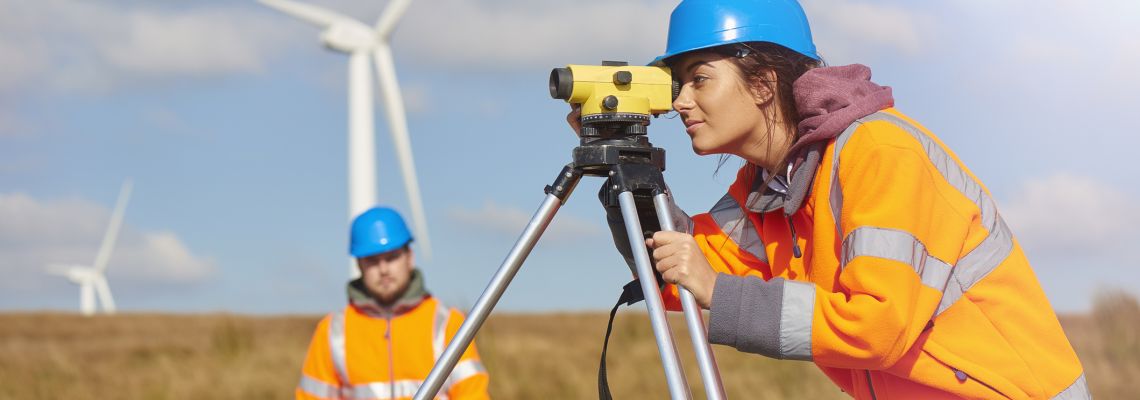 two wind farm engineers using a builder's level to plan out the expansion of the wind farm site. they are wearing orange hi vis jackets and blue hard hats . one is male , one is female. In the foreground the female is looking through the level whilst the male engineer is approaching .In the background wind turbines can be seen across the landscape.
