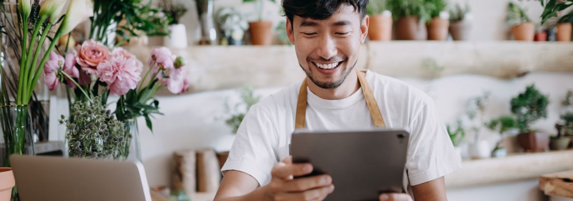 Asian male florist, owner of small business flower shop, using digital tablet while working on laptop against flowers and plants. Checking stocks, taking customer orders, selling products online. Daily routine of running a small business with technology Asian man uses bachelors in business to start small business.