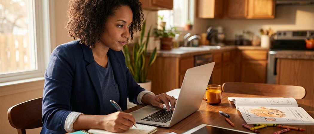Mid-career learner studying at a kitchen table at night with a laptop, notepad, and a child's homework nearby, lit by warm window light to convey focus, balance, and progress.