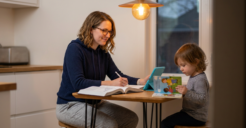 Adult learner studying at a kitchen table in the evening, balancing a textbook and tablet while a child reads nearby, lit by soft window and pendant light to convey family and study balance.