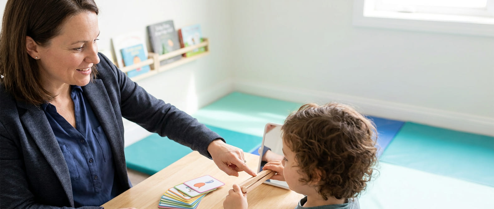 Speech-language pathology assistant works with young child at table, using picture cards and a tablet during therapy session in a bright classroom, illustrating supportive communication practice and pediatric care.