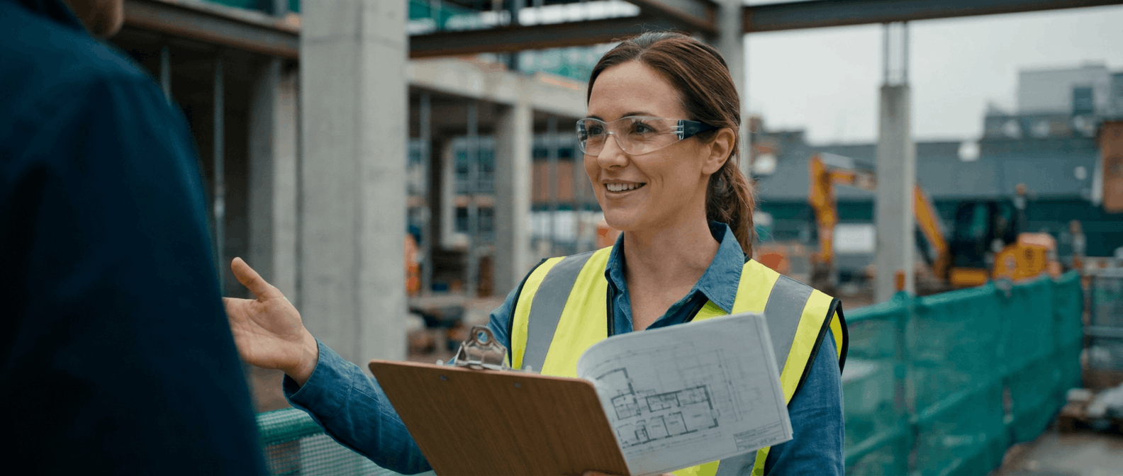 Construction manager reviewing plans on an active jobsite with a commercial build in the background.